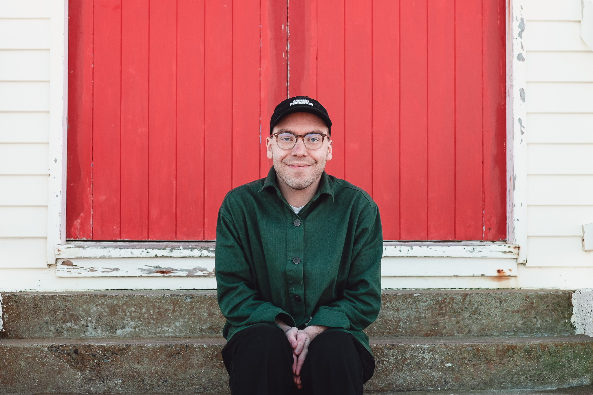 Our Artists Image Jesse Austin-Stewart sits in front of a red-painted wooden door smiling with his hands clasped in-front of him.