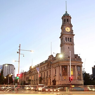 Auckland Town Hall's iconic clock tower is softly lit at dusk with street lights illuminating the foreground and the sky a light purple shade.  Image credit: Auckland Live